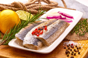 Atlantic Marinated herring fillet with pepper, tomatoes, herbs, onion and lemon on white plate on cutting board.