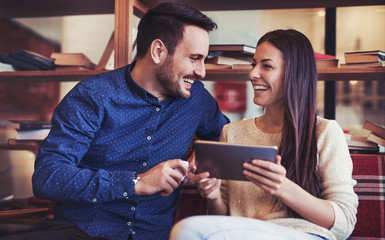 Young couple sitting in the library and having fun with tablet, surrounded with books on the wooden shelves