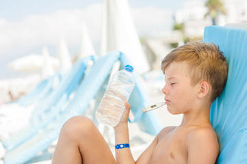 Sad little boy on hot beach with sunstroke measures the temperature and holds a bottle of water