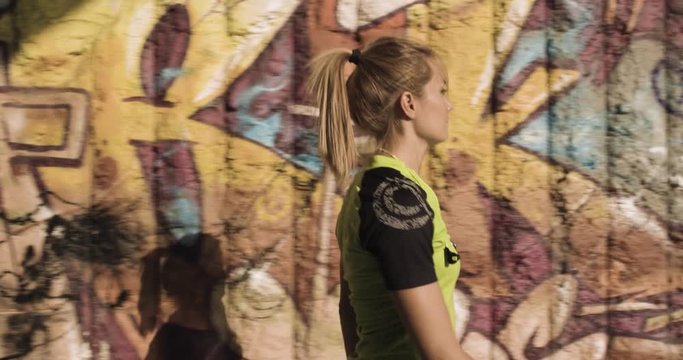 Young blond woman in fitness wear walking near grunge graffiti wall.Side following view.Summer sunny day.Industrial green city.Urban runner cardio healthy activity workout.4k slow motion 60p video