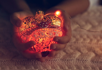The child holds a glass heart, a Christmas toy - a garland in the hands on the background of a warm knitted sweater. Night