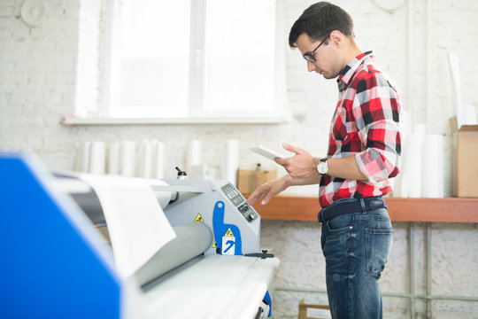 Side view of casual man with tablet working with printing machine in modern publishing office
