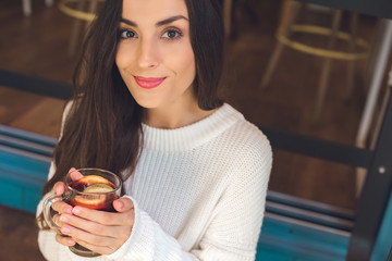 smiling young woman looking at camera and holding cup of mulled wine at table in cafe