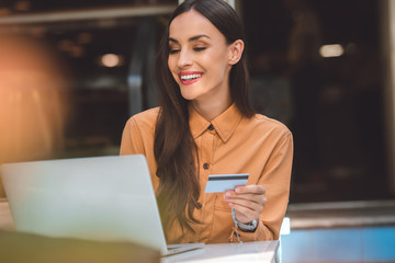 cheerful stylish woman with credit card doing online shopping on laptop at table in cafe at city street