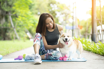 A healthy woman playing with a corgi puppy while exercising on yoga mat surrounding with gym tools such as kettlebell and dumbbell, outdoor training with dog