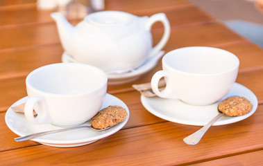 Two white cups and spoons with biscuits, teapot on a wooden table