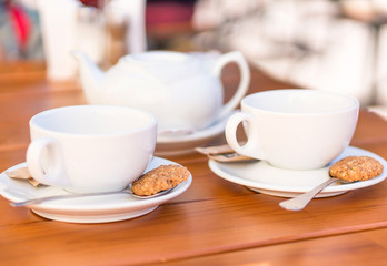 Two white cups of tea and spoons with biscuits, teapot on a wooden table