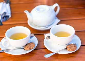 Two white cups of tea and spoons with biscuits, teapot on a wooden table