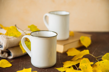 two white mugs surrounded by yellow leaves with a book and a knitted scarf, the concept of warmth and autumn sadness