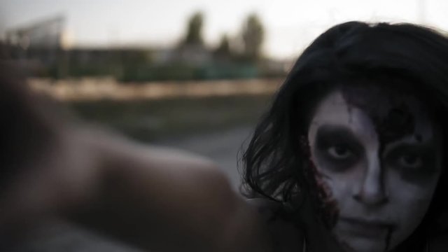 Portrait of a creepy zombie woman with wounded face and clawing hands walking outdoors with an industrial abandoned place on the background. Halloween, filming, creepy concept