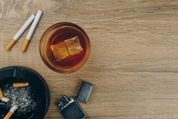 Fototapeta na wymiar Top view of a glass of bourbon whiskey, cigarettes, metallic lighter and black ceramic ashtray full of ashes with the cigarette butts on the wooden table
