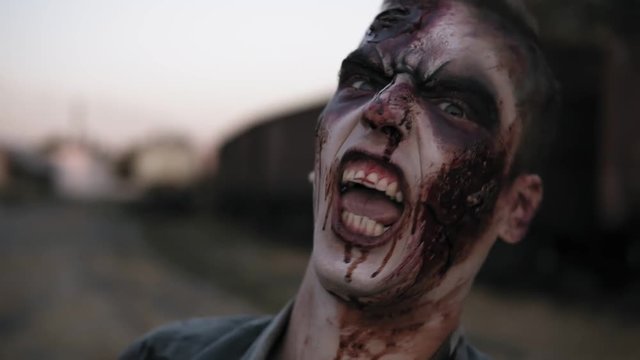 Portrait of a male zombie with bloody teeth and wounded face screaming and shouting. Halloween, filming, staging concept. Blurred railway wagons on the background