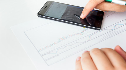 Closeup image of business analyst using smartphone calculator while analyzing stocks sales graph