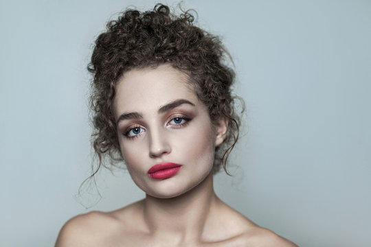 Beauty portrait of young beautiful naked fashion model with collected dark curly hair, nude makeup and red lips looking at camera with serious face. indoor studio shot. isolated on gray background.