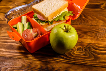 Bottle of water, green apple and lunch box with sandwich, cucumbers and tomatoes on wooden table