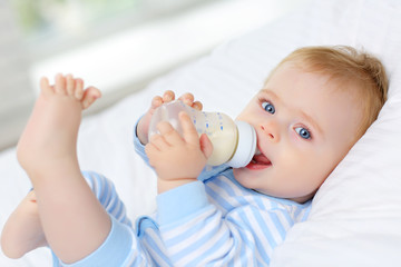 Charming blue-eyed baby 7 month old lies in bed and drinks milk from a bottle - obrazy, fototapety, plakaty
