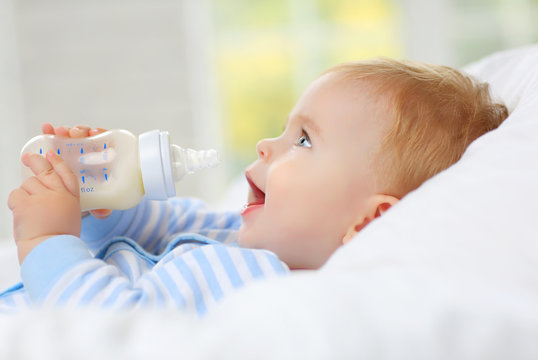 Charming blue-eyed baby 7 month old lies in bed and drinks milk from a bottle