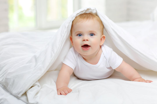 Charming blue-eyed baby 7 month old lies in bed in a white bodysuit, covered with a blanket and smiles