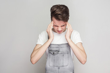 Portrait of young man suffering from severe headache, pressing fingers to temples, closing eyes to relieve pain with helpless face expression. Isolated, studio shot, copy space, on grey background