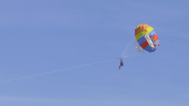 Couple of unrecognizable people are flying on a multi-colored parachute in the sky, which is tied to a motor boat. Parasailing in clear sunny weather
