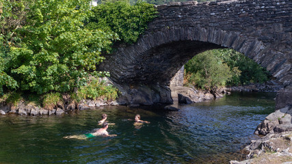 Fototapeta na wymiar Father and teen children swimming in the River Duddon by old stone bridge in Ulpha in the Lake District National Park, UK. Scenic view of English countryside on a sunny summer day.