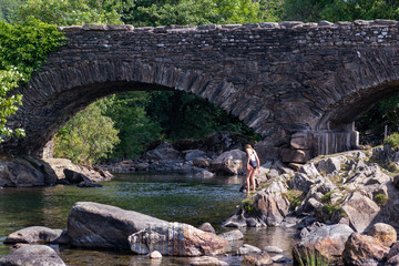 Fototapeta na wymiar Teenage girl is entering into the River Duddon by old stone bridge in Ulpha in the Lake District National Park, UK. Scenic view of English countryside on a sunny summer day.