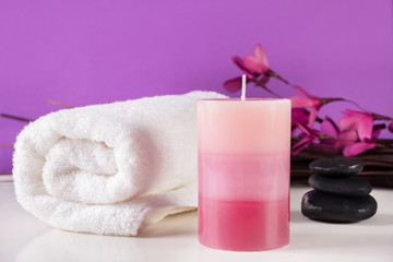 Fototapeta na wymiar Aromatic decorative candle, white towel and black stones and purple flower on branches in background. Spa treatment concept