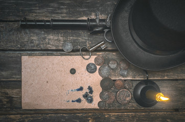 Blank page secret letter, weapon, money, bowler hat, rusty key and burning candle on the wooden desk table. Retro style detective agent table.
