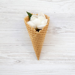 Sweet waffle cone with white peony flowers on a white wooden background, top view.