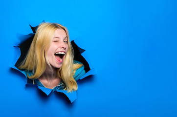 View of woman face through hole in blue paper. Winking smiling girl making hole in paper. Fashion and beauty concept. Copy space for advertising, to insert text or slogan. Discount, sale, season sales