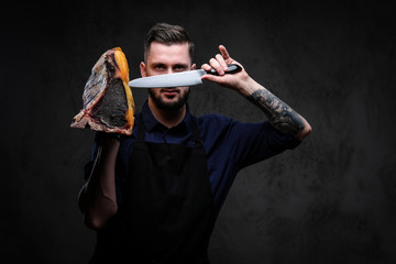 Chef cook holds the knife and large piece of exclusive cured meat on a dark background.