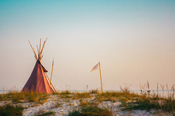 Red wigwam and white flag on the sandy seashore. Indian native national building outdoors summer. Teepees on coast. © baklykovadaria