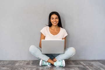 Happy young woman sitting on the floor with crossed legs and using laptop on gray background.