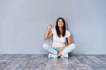 Have idea! Cute woman in casual clothes sitting with legs crossed on the floor gesturing index...