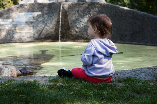The little girl sits on the shore of the pond, next to the fountain, hold a stick like a fishing rod and looks at the water. Back view. Child playing with water of city fountain. Lifestyle concept