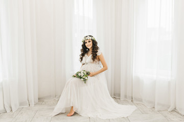 Attractive young pregnant girl in a white dress with curls, a wreath and a bouquet of flowers. Photo session waiting for the baby