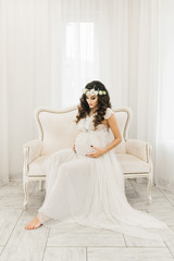 Fototapeta premium Attractive young pregnant girl in white dress with curls. Photo session waiting for the baby