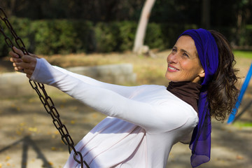 Happy beautiful woman with purple shawl on head swinging in the playground on sunny day. Care free...