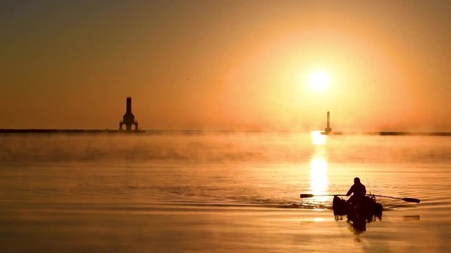 A lone man rows his boat in the golden sunlight of sunrise. Featuring the Port Washington WI lighthouse, breakwater, sea fog, lake michigan