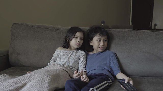 Kids shocked by something they see on TV while watching it and sitting on sofa together in mostly barren minimalist room.