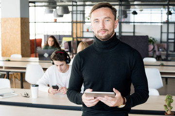 Waist up portrait of handsome contemporary businessman looking at camera and holding digital tablet while posing in open space office of startup company, copy space