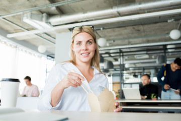 Portrait of cheerful blonde woman eating Chinese food at workplace and smiling at camera, copy space