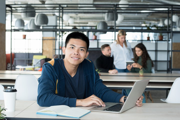 Portrait of young Asian man smiling at camera while working as intern in open space office of IT company