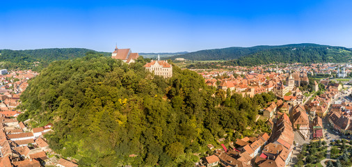 Aerial panorama of medieval Sighisoara in Romania with blue sky, red roofs, bastions, towers and city walls above the Mures river