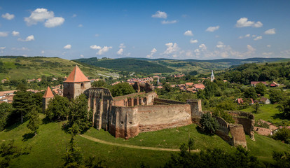 Aerial view of ruined Gothic Saxon medieval Slimnic castle near Sibiu, Romania with donjon, church, barbican, walls on a green hill with blue cloudy sky