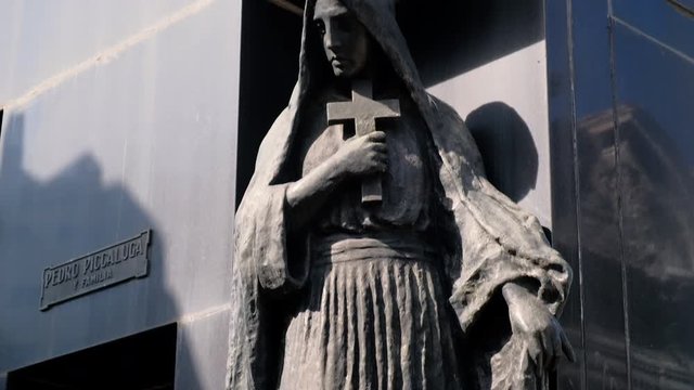 Woman holding cross made of black stone statue in La Recoleta Cementery at daytime long shot tilt down to high