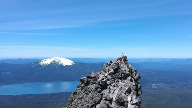 Man walking up to the top of a cliffside mountain with beautiful lake and volcano in the background. Aerial shot.