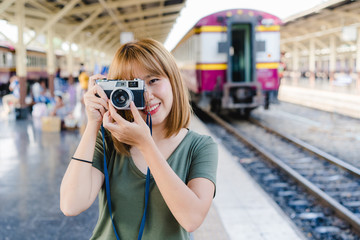 Obraz premium Cheerful Traveler backpacker Asian woman using camera to make photo while travel at train station in Bangkok, Thailand. Woman tourist journey on holidays concept.