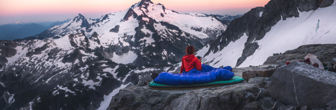 Girl in sleeing bag drinking from mug infront of large glacier covered alpine peak
