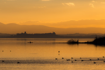 Naklejka na ściany i meble Beautiful view of Trasimeno lake (Umbria, Italy) at sunset, with orange tones, birds on water, a man on a canoe and Castiglione del Lago town on the background